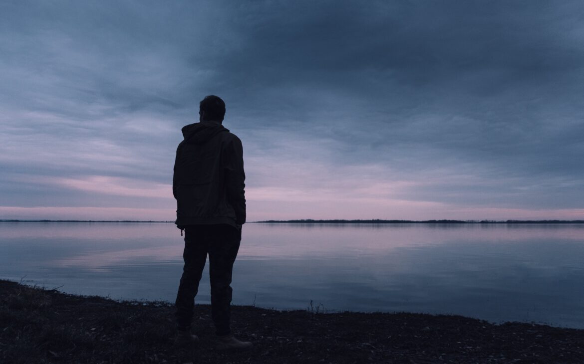 Silhoutte photo of a man standing next to a water body, clear view of the sky